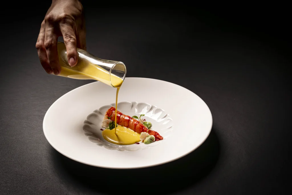 Una mano vierte salsa amarilla sobre una elegante langosta en un plato blanco, adornada con hierbas. El fondo oscuro resalta la sofisticada presentación del plato.