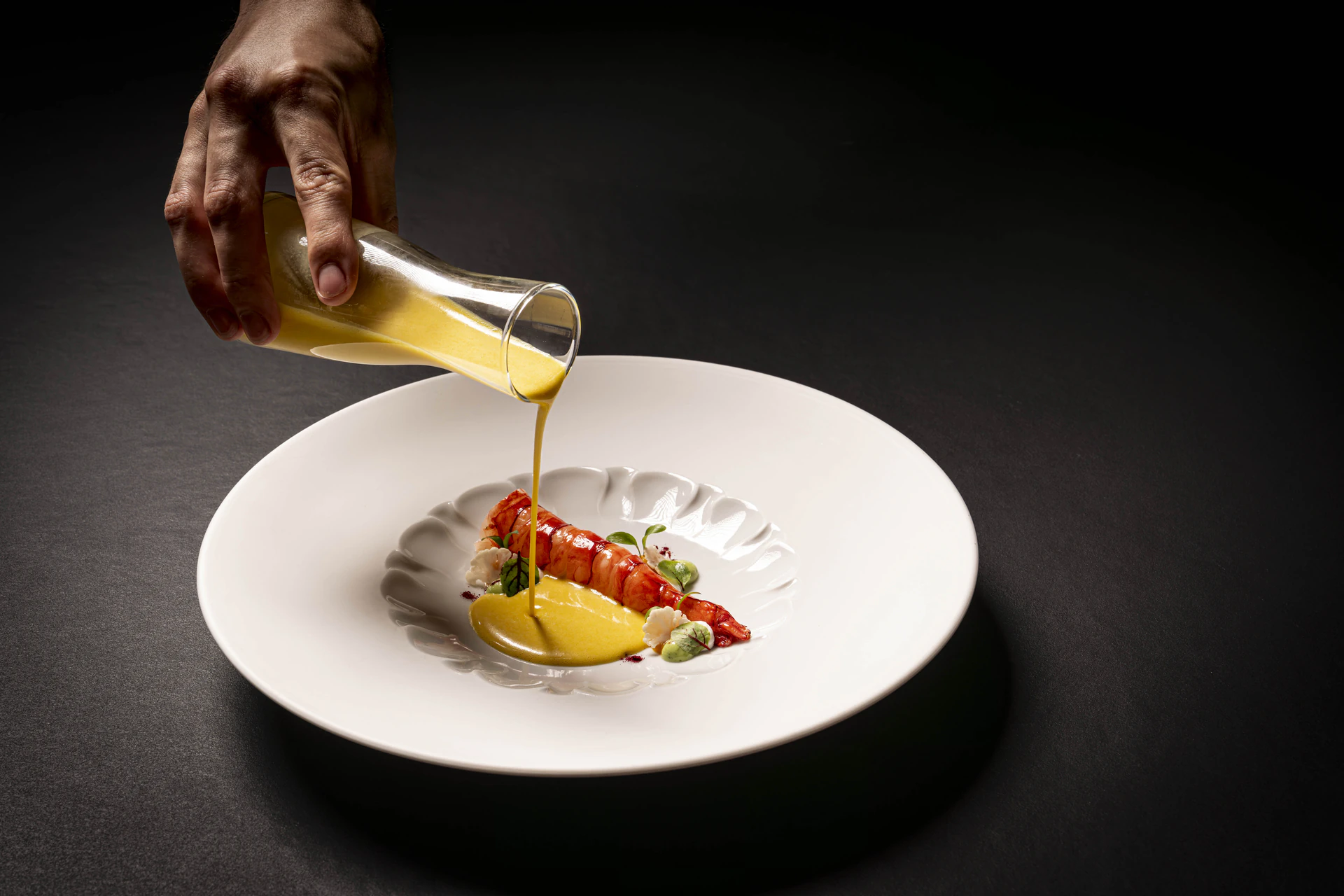 Una mano vierte salsa amarilla sobre una elegante langosta en un plato blanco, adornada con hierbas. El fondo oscuro resalta la sofisticada presentación del plato en un evento gastronómico.